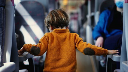 Young boy in a sunny yellow pullover navigates the train corridor, hands grasping the seat...