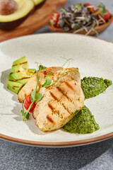 Macro vertical side view capturing the textures of grilled chicken fillet, avocado, and spinach on a ceramic plate. Set against a grey concrete background with hard shadows