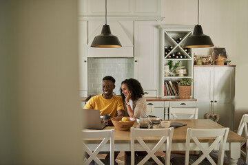 Smiling young multiethnic couple using a laptop during breakfast at home - Powered by Adobe