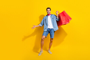 Full size photo of satisfied cheerful guy wear jeans jacket holding shopping bags buy new clothes isolated on yellow color background