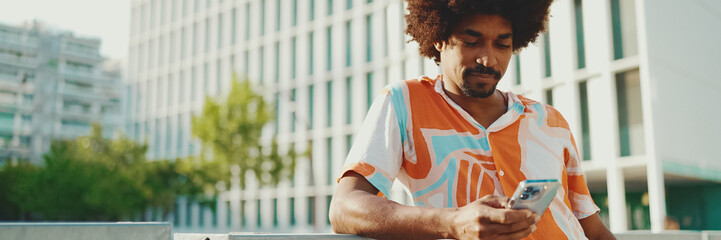 Closeup young African American man wearing shirt writes in social networks on mobile phone on urban...