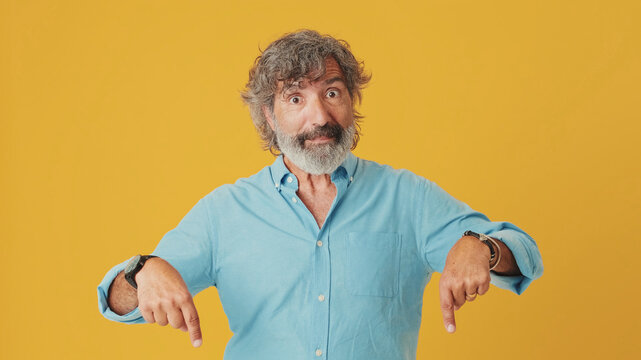 Elderly grey-haired bearded man wears blue shirt, pointing fingers down on workspace area copy space mock up, isolated on orange background in studio