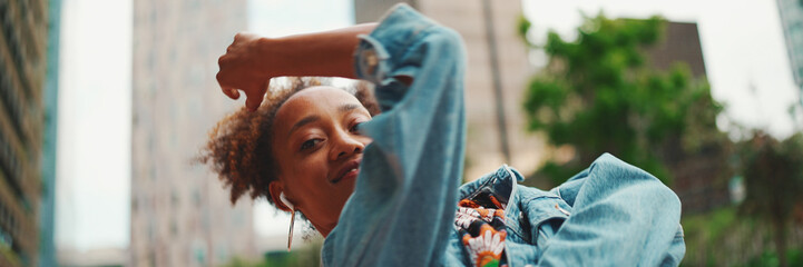 Closeup, smiling African girl with ponytail wearing denim jacket, in crop top with national pattern listening to music on headphones and dancing outdoors.