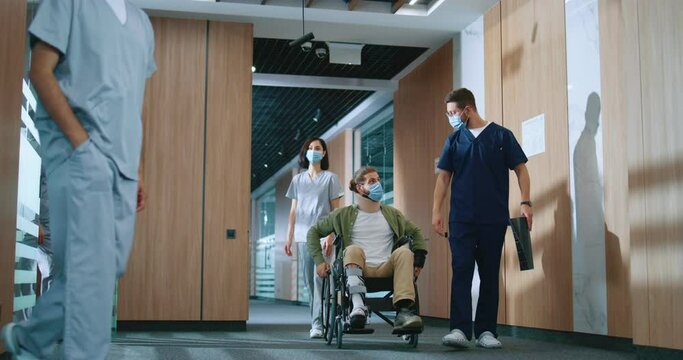 Injured Caucasian male patient in wheelchair moving around hospital corridors with professional helpful doctor in uniform holding x-ray talking and consulting man. Modern clinic concept.