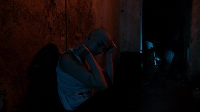 A bald woman who is a drug addict feels withdrawal symptoms without drugs. Old basement in neon light. Hopeless drug addict