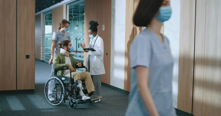 Young Caucasian bearded man sitting in wheelchair and talking with helpful medical staff in modern hospital. Beautiful successful nurse and female African-American doctor consulting patient.