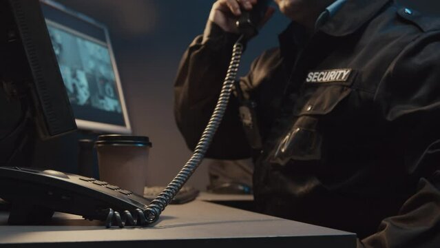 Midsection cropped shot of unrecognizable security officer answering work call on landline phone while monitoring CCTV video footages in guard booth