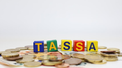 Singola parola "TASSA" su blocco di legno, tasse isolate, con sfondo di monete in euro. © barbacane