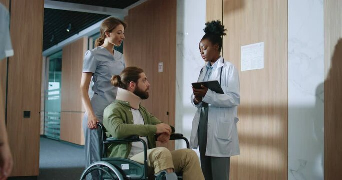 Young Caucasian nurse and professional African-American doctor talking and helping injured young man in wheelchair recovering in hospital. Experienced physiotherapist consulting patient.