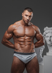 Fototapeta premium Handsome man exuding a rugged charm, with a flawless muscular chest, posing proudly beside ancient Greek statues against a gray backdrop