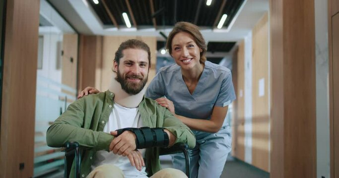 Portrait of happy injured Caucasian man recovering in hospital with qualified female doctor looking at camera and smiling. Bearded young patient and helpful nurse in modern clinic.