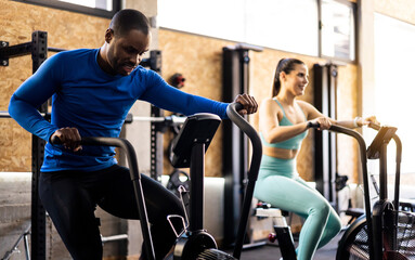 A couple of different ethnicities are doing functional training with stationary air bikes.African...