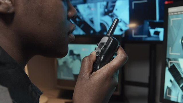 Closeup side shot of security officer talking on handheld transceiver while monitoring CCTV video footages on multiple monitors in surveillance room