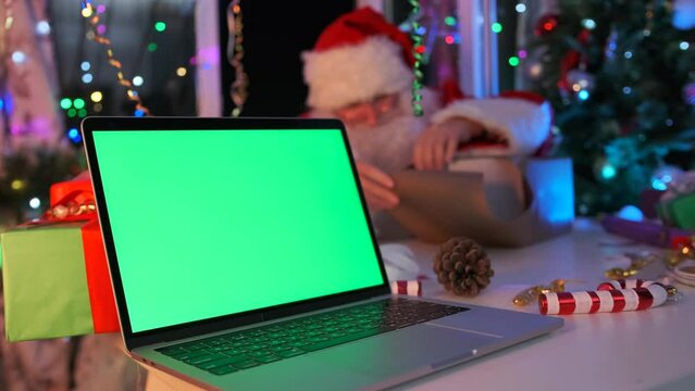 Laptop with green chromakey screen lies on table next to Santa Claus packing box with gift in wrapping paper in background among New Year decorations,  tinsel, Christmas Tree with glowing garlands.