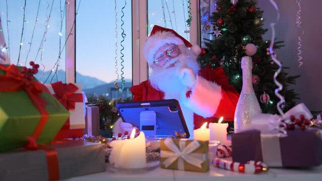 Grandfather Santa sits with tablet communicates and wishes children Merry Christmas, among Christmas decorations of burning candles, gift boxes, tinsel and garlands, with xmas tree on background. 