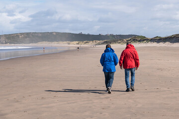A couple in their colorful coats walks along the beach. Salinas Beach, Asturias