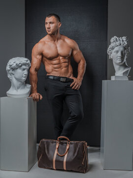 Man with a well-groomed model appearance with a bare muscular torso, posing with a luxurious high-fashion bag next to ancient Greek statues on a gray background