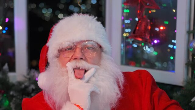Middle age man wearing Santa Claus costume and beard shows an obscene gesture by putting his fingers to his mouth and sticking out his tongue, with festively decorated room at Christmas party.