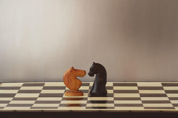 Two chess knights, one wooden and one marble, stand facing each other on a chessboard. The...