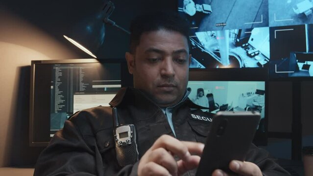 Chest up shot of bored Biracial security man in uniform with walkie-talkie using smartphone during workday in surveillance room with multiple monitors