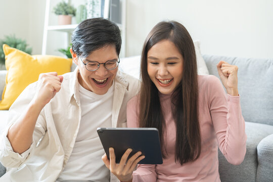 Happy excited, smiling asian young couple love using tablet pc, great deal or business success, received or getting cash back, tax refund, good news by mail while sitting on floor at home.