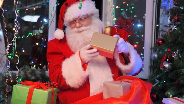 Funny Santa collects beautiful gift boxes into red present bag, dancing and shaking them, sitting against background of Christmas glow garlands and shiny tinsel at night Xmas party. New Year. Close up