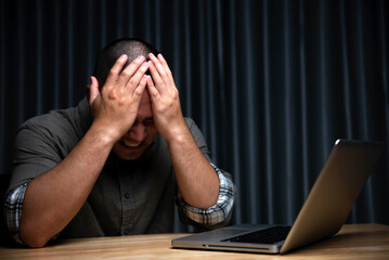 A man holds his head with both hands in stress on his desk and a laptop