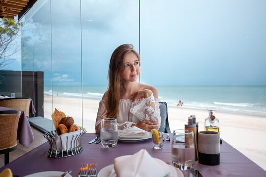 Young woman enjoys upscale dining experience with ocean view, fine bread selection on table, serene beach backdrop. Upscale travel and dining