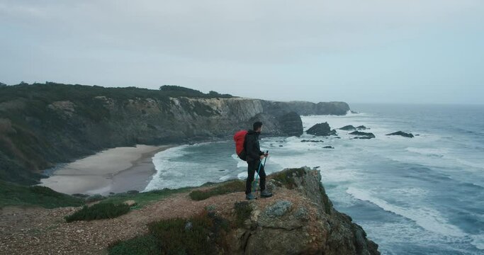 Wide open shot of young hispanic man look into distance over ocean and cliffs, stand on edge of rock on end point of pilgrim trail or path. Hiking and exploring wild coastline