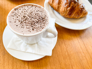 Hot Milk Chocolate Drink in white porcelain mug next to croissant on a wooden table. Concept for breakfast, working, task, job, business plan. Empty blank copy text space.