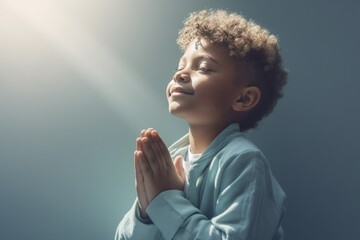 Little black boy on knees holding hands and praying in the morning, pastel neutral background. Christianity, faith, spirituality, religion, salvation, peace, faith concept. Kid praying to God - obrazy, fototapety, plakaty