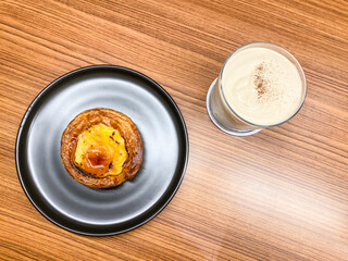 Hot Cappuccino Coffee with latte art in a glass next to egg tart on wooden table. Caffe Latte or Coffee Milk Drinks and bread. Concept for relaxing time, working and business.