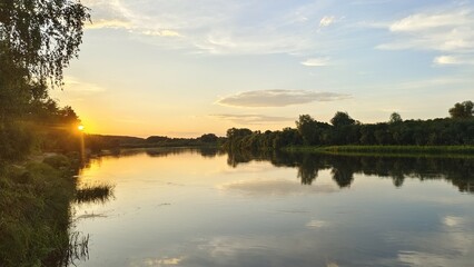 On a summer evening, the sun among the trees sinks below the horizon. The colorful cloudy sky is reflected in the river water. There are trees growing on the grassy banks and calamus in the water