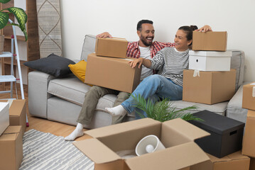 Positive couple of adult man and woman rejoice at new purchase of apartment sitting on sofa with stacks of packed boxes with things for moving in living room