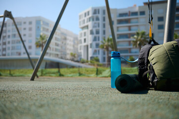 Sports tools on the sports ground. A backpack, bottle of water and rolled fitness mat. Sport....