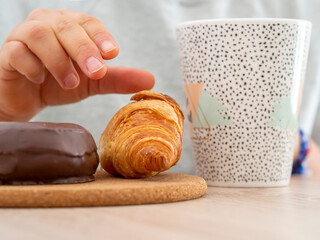 Close-up of an unrecognizable woman hand grabbing a croissant for a sugary snack with a coffee mug....