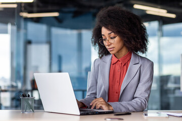 A young African-American businesswoman is sitting at a desk in the office and is concentrating on working on a laptop.