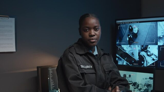 Portrait of Black female guard using handheld transceiver and then looking at camera standing in surveillance room with multiple screens of CCTV video footage