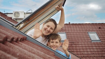 Smiling mother and her son open attic windows, gaze outside, and warmly wave towards the camera....