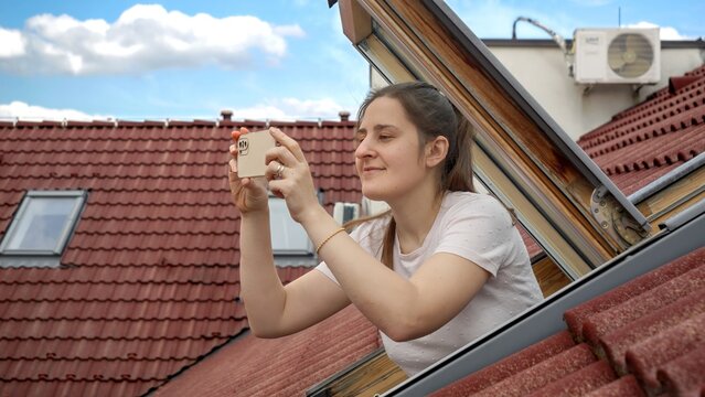 Young woman, with a bright smile, uses her smartphone to capture photos while looking out of an open attic window. Tourism, holidaying in Europe, and the spirit of travel and exploration