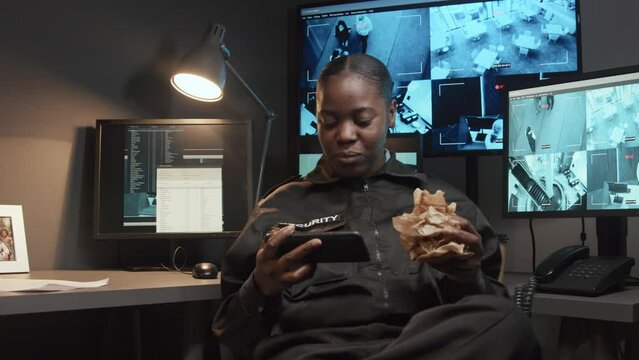 Medium shot of young careless African American security officer eating snack and watching video on phone not monitoring CCTV footage in guard room
