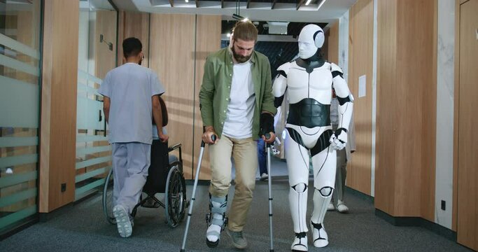 Young beautiful injured patient struggling to walk with crutches and helpful robot assistant in future hospital helping. Artificially intelligent white bot working at clinic assisting people.