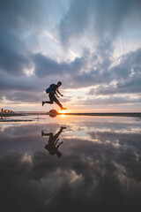Fototapeta premium Brown-haired man with a backpack, aged 25-29, jumps into a pool of water on the beach during sunset. Catching the sun between the ground and his feet. Oostende, Belgium