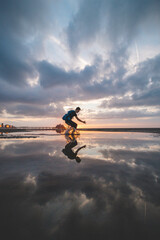 Brown-haired man with a backpack, aged 25-29, jumps into a pool of water on the beach during sunset. Catching the sun between the ground and his feet. Oostende, Belgium