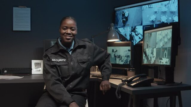 Portrait of young smiling African American guard in black uniform controlling CCTV video footage and then looking at camera sitting in surveillance room