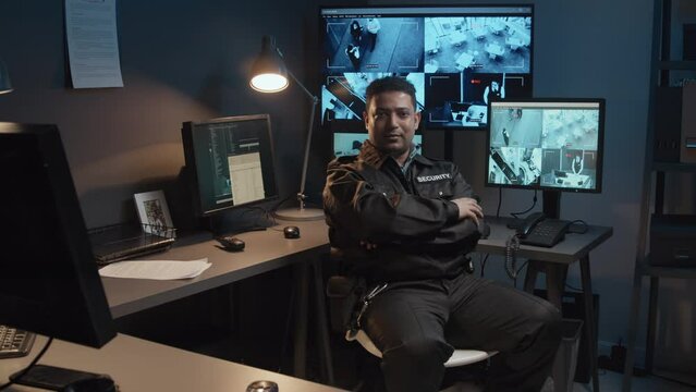 Portrait of smiling male Biracial security officer controlling surveillance CCTV video footage on multiple monitors and then turning to camera in guard room