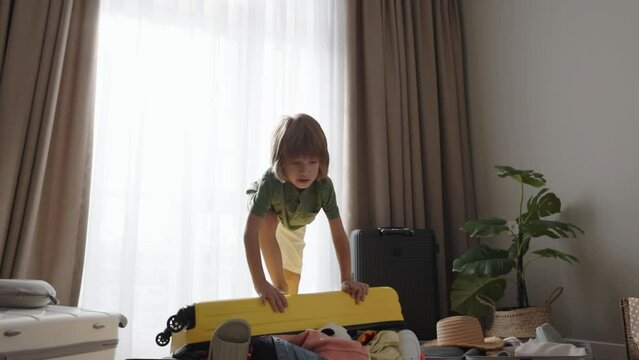 Boy throws another toy into his overfilled and disorderly suitcase. Then he climbs onto it with his feet and jumps on it, trying to close. Vacation dreams chaotic travel preparations, app advertising