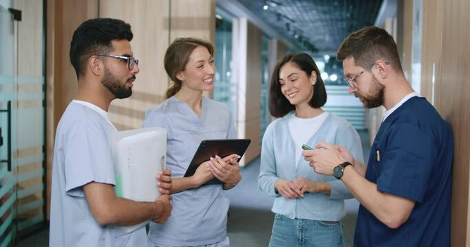 Side view portrait of diverse group of young skilled medical professionals chatting in modern hospital corridor during break. In uniform successful medical workers conversing and having fun.
