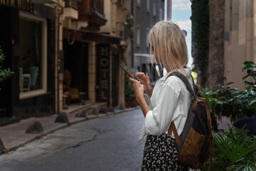 Young blonde female tourist in a white shirt, skirt, with a craft backpack stands with a mobile phone in her hands on a beautiful street in Istanbul. Concept of tourism, modern technologies.