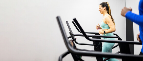 A young couple of different ethnicities is running on a treadmill inside a gym. The young woman is...